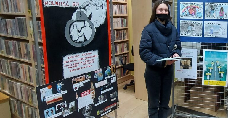 Zdjęcie przedstawia laureatkę pierwszego miejsca w konkursie plastycznym Zofie Czader . w tle ekspozycja nagrodzonych prac plastycznych